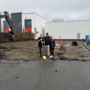 Three people stand together at a construction site on a wet day, holding a golden shovel. Behind them are a wooden fence, a white building, an excavator, and some construction cones and equipment.