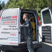 A technician stands at the open back doors of a white Gandy Installations van, organizing tools and yellow cables for a boiler repair. The van displays HVAC service details and contact information. Trees and a sidewalk are visible in the background.