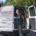 A technician stands at the open back doors of a white Gandy Installations van, organizing tools and yellow cables for a boiler repair. The van displays HVAC service details and contact information. Trees and a sidewalk are visible in the background.