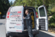 A technician stands at the open back doors of a white Gandy Installations van, organizing tools and yellow cables for a boiler repair. The van displays HVAC service details and contact information. Trees and a sidewalk are visible in the background.