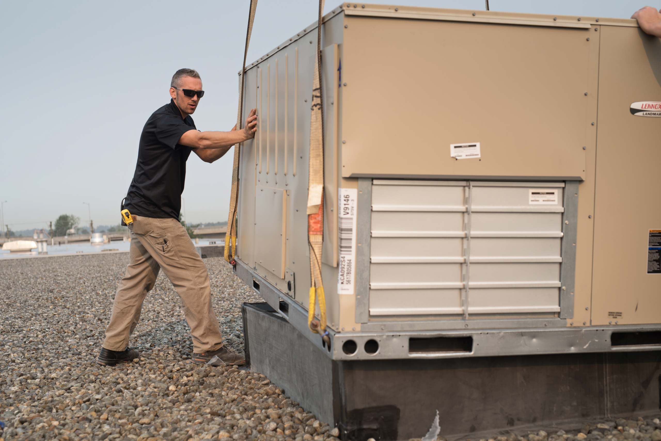 A man in sunglasses and work clothes guides a large industrial HVAC unit, including ceiling unit heaters, as it is lifted by straps on a gravel-covered rooftop.