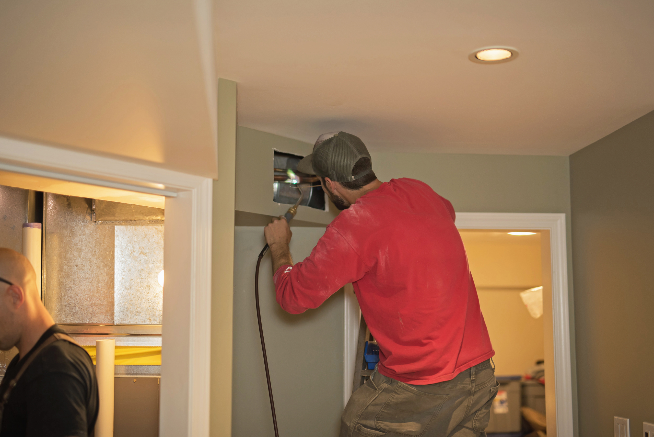 A man in a red shirt and cap repairs an air vent in a wall, possibly checking connections to furnaces, while standing on a step stool inside a home. Another person is partially visible nearby, working in a different room.