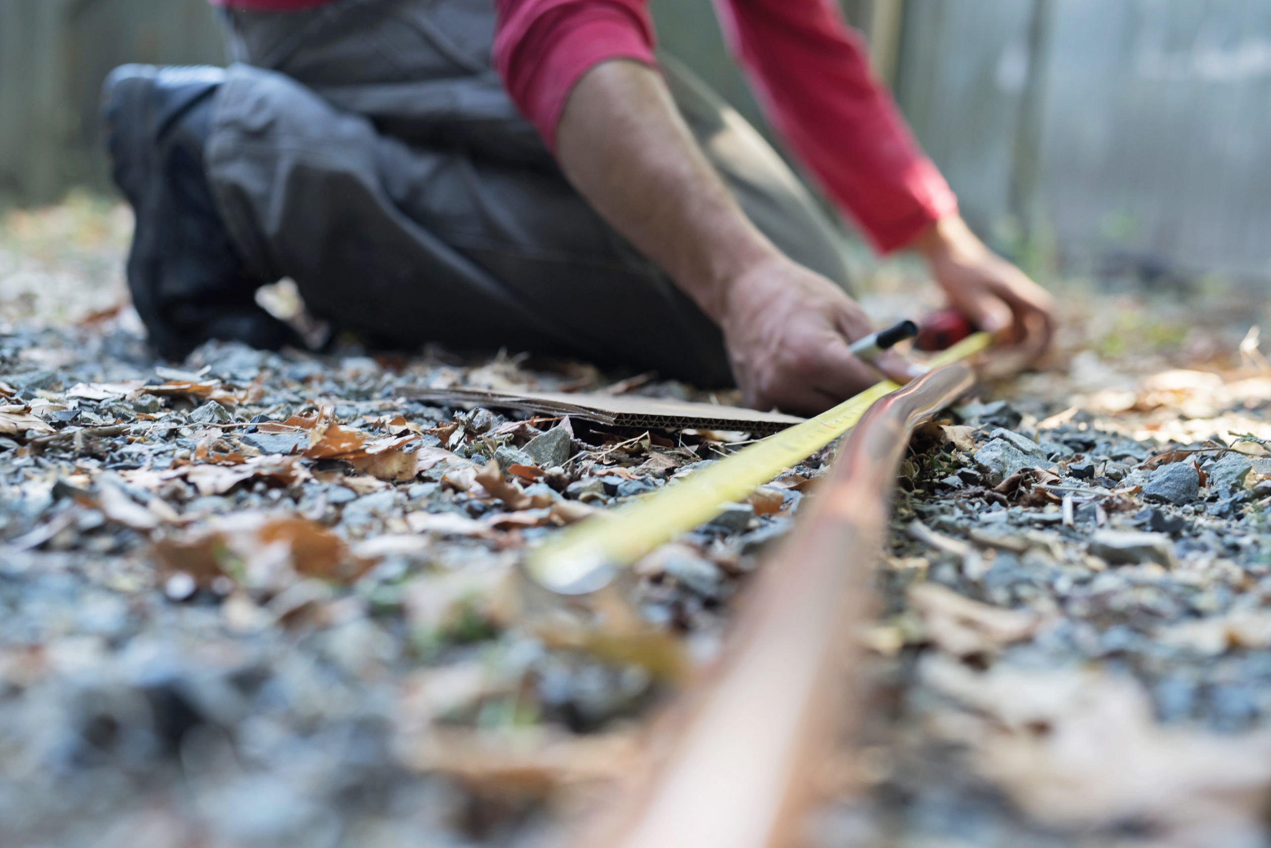 A person kneels on the ground, using a measuring tape and a ruler to measure along a copper pipe near furnaces, surrounded by gravel and fallen leaves outdoors.