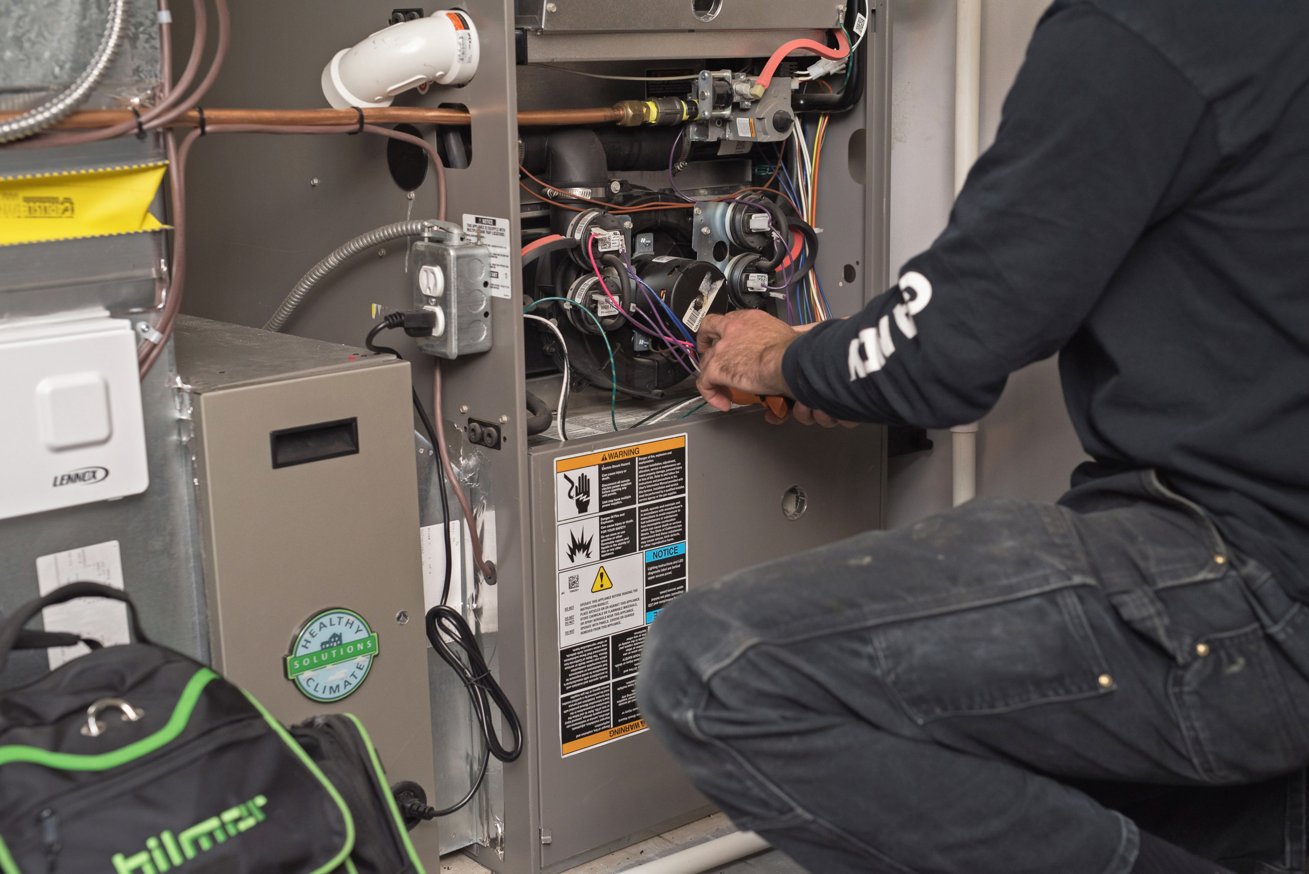 A technician is kneeling and repairing the wiring inside an open furnaces unit, surrounded by tools and equipment. The technician's face is not visible.