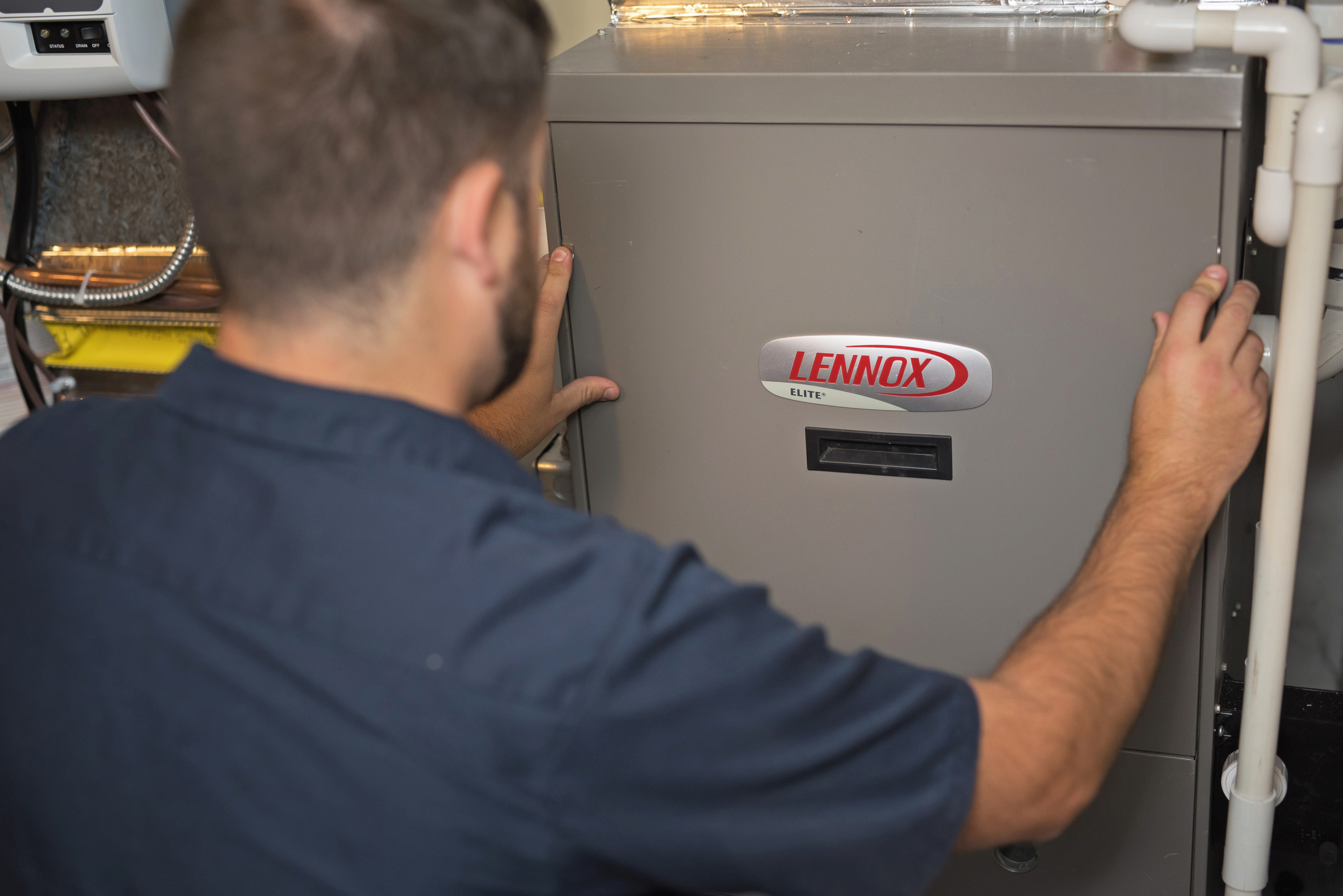 A person in a navy shirt is inspecting or repairing a Lennox Elite HVAC furnace unit, possibly preparing for a furnace replacement. They use both hands to check the front panel, with pipes and wires visible nearby.