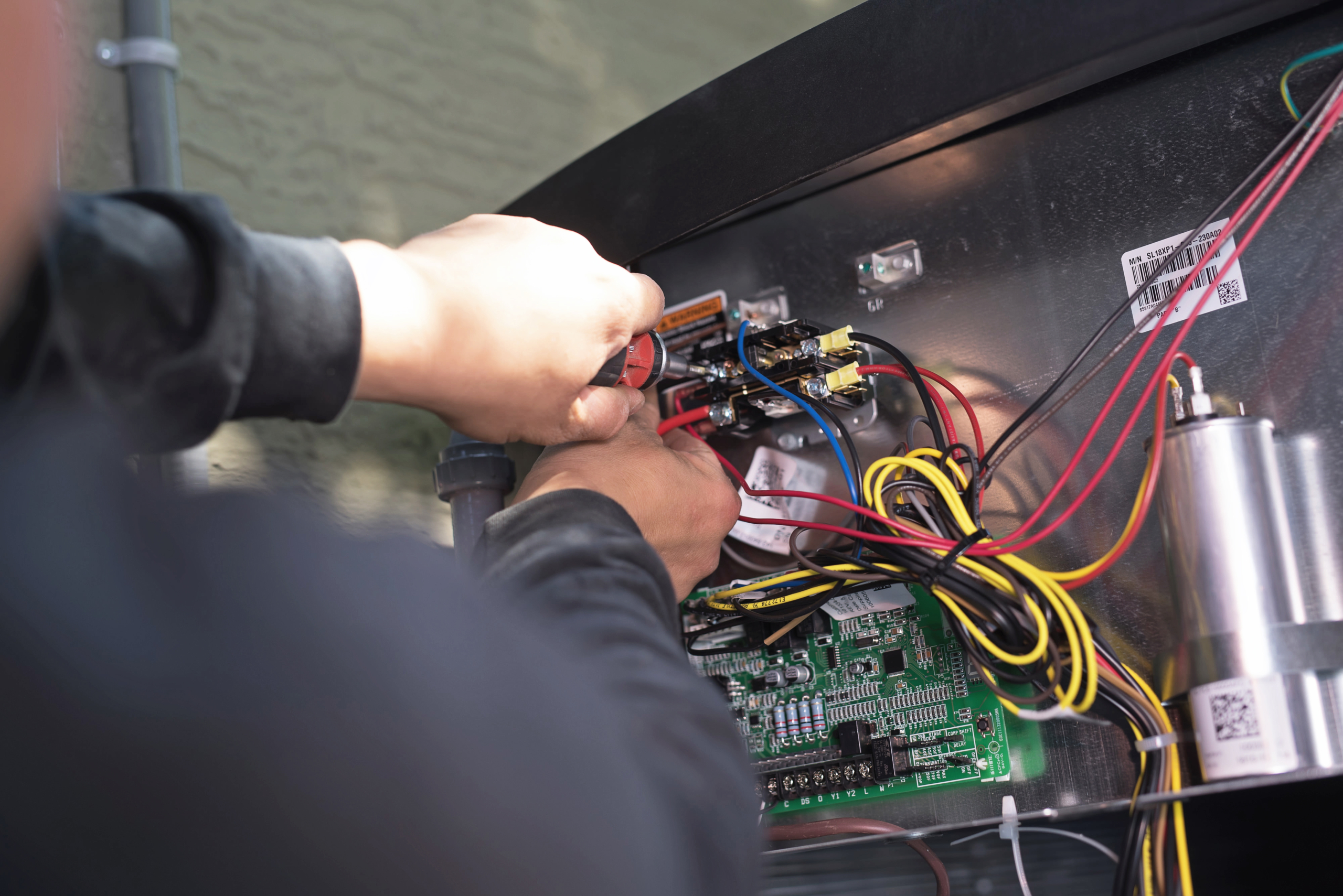 A person performs heat pump maintenance, using a screwdriver to work on the wiring and electronic components inside an open control panel with various colored wires and circuit boards visible.