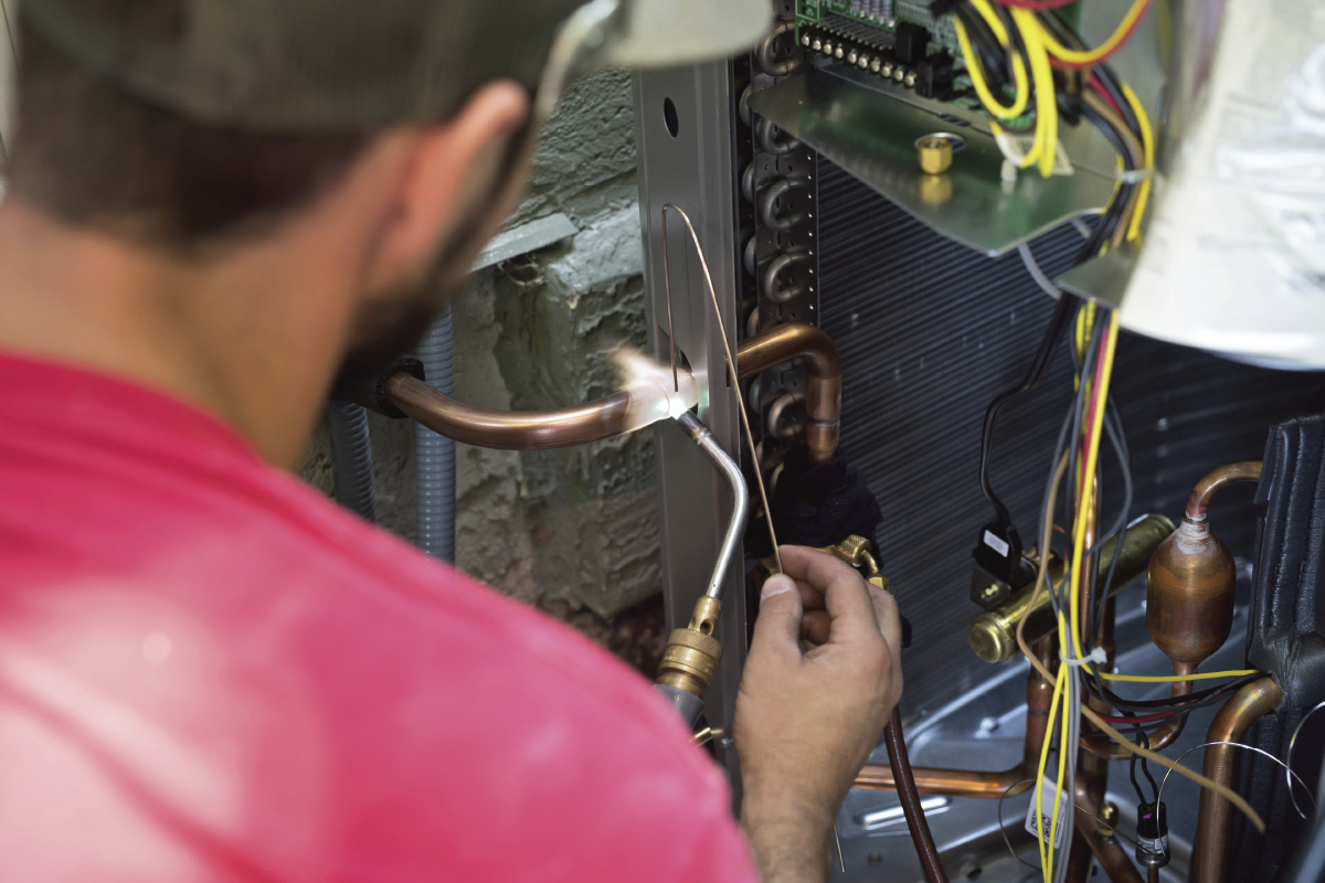 A person in a red shirt uses a torch to solder copper pipes inside an HVAC unit, surrounded by exposed wires and circuit boards—showcasing detailed heat pump maintenance or repair work on air conditioning equipment.