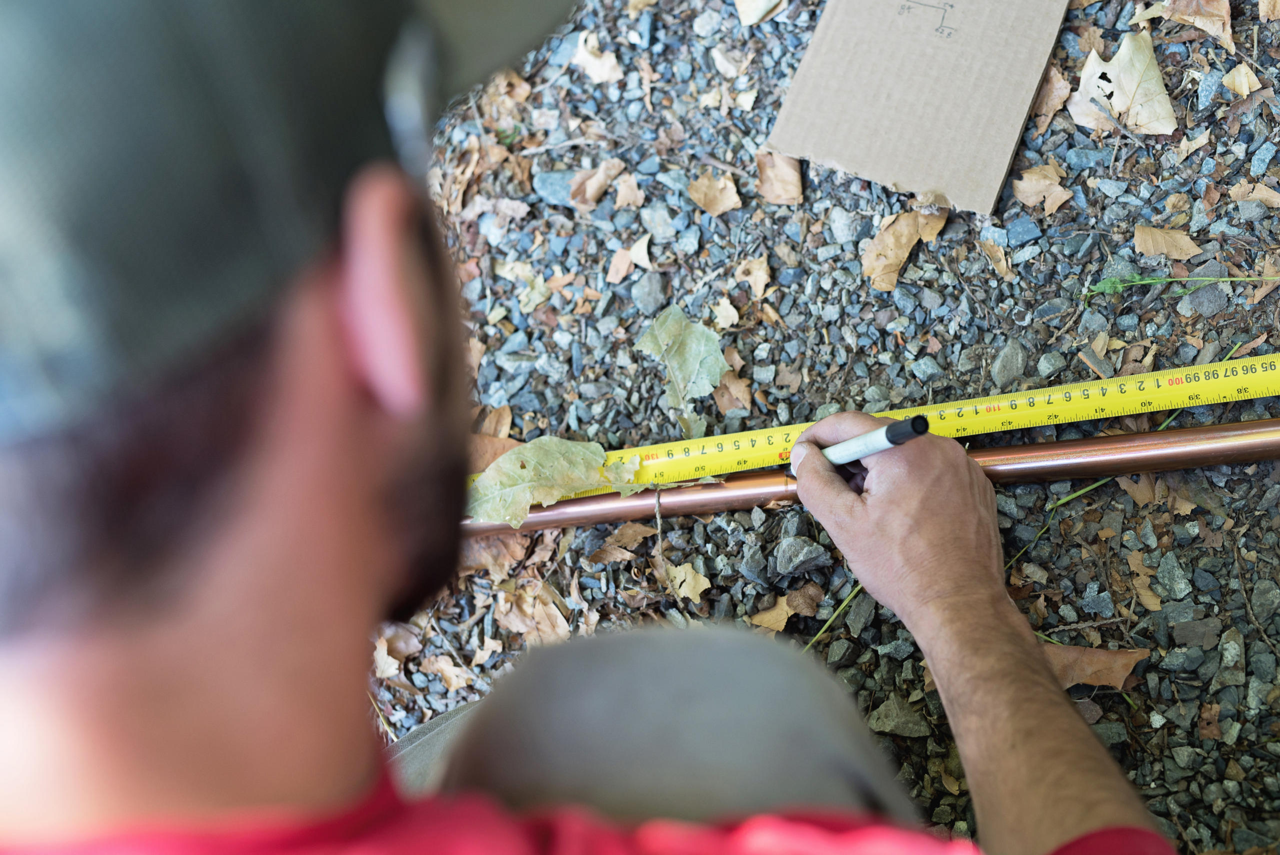 A person in a red shirt and cap measures a copper pipe for ductless mini split installation with a yellow tape measure and marks it with a black marker while kneeling on gravel and fallen leaves outdoors.