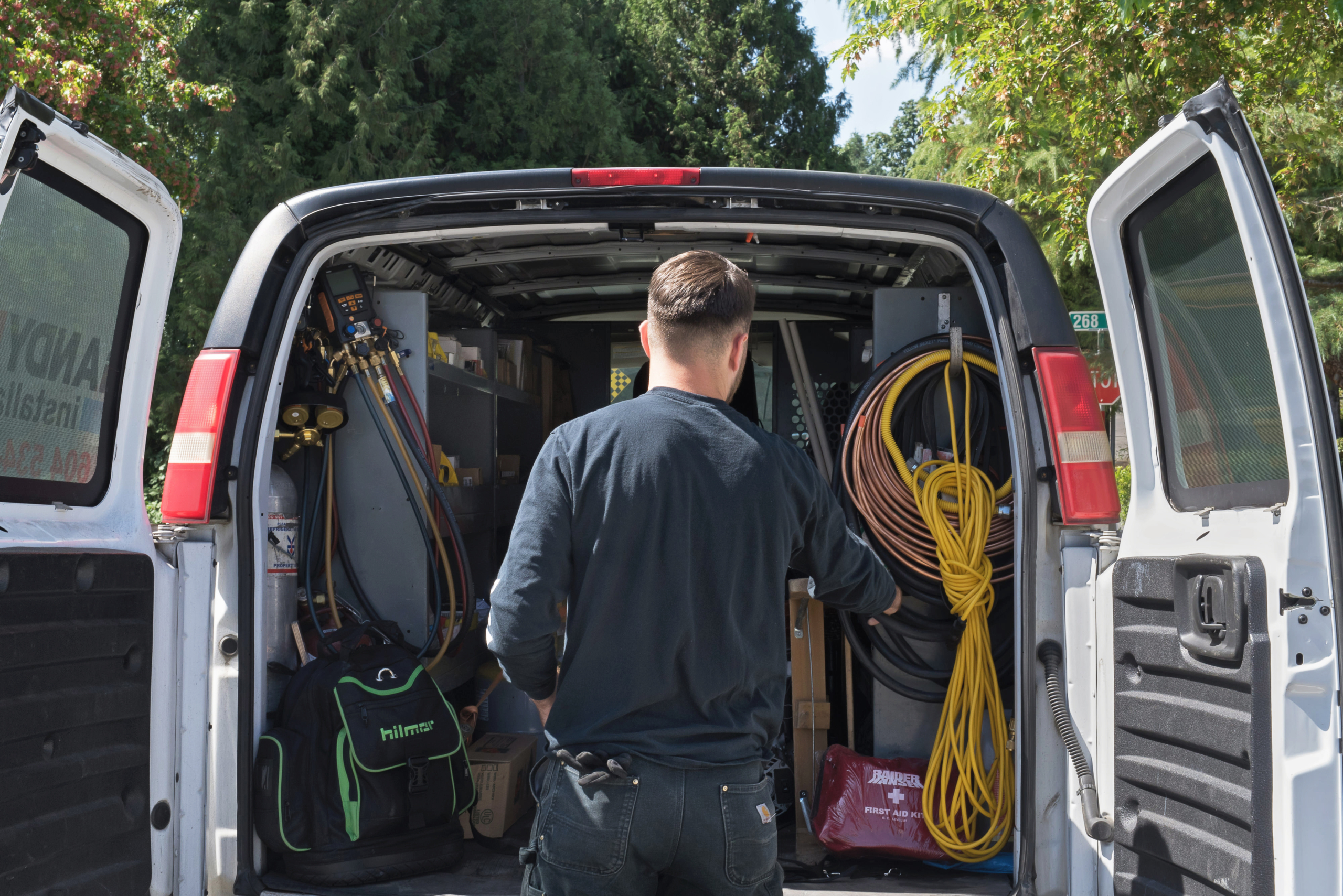 A man stands at the open back of a work van, facing inside. The van is organized with various tools, cables, and ductless mini split maintenance equipment, surrounded by trees and greenery outside.