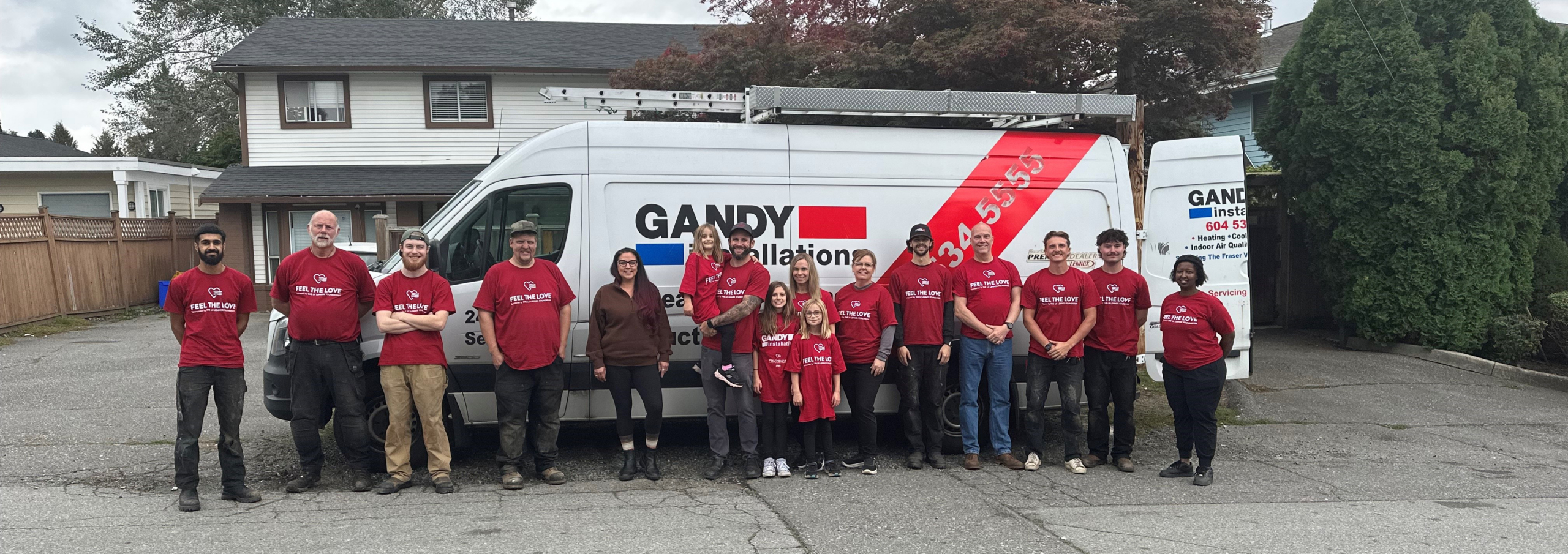 A group of 13 people in matching red shirts stand in front of a white Gandy Installations van parked in a residential driveway, with houses and trees in the background.