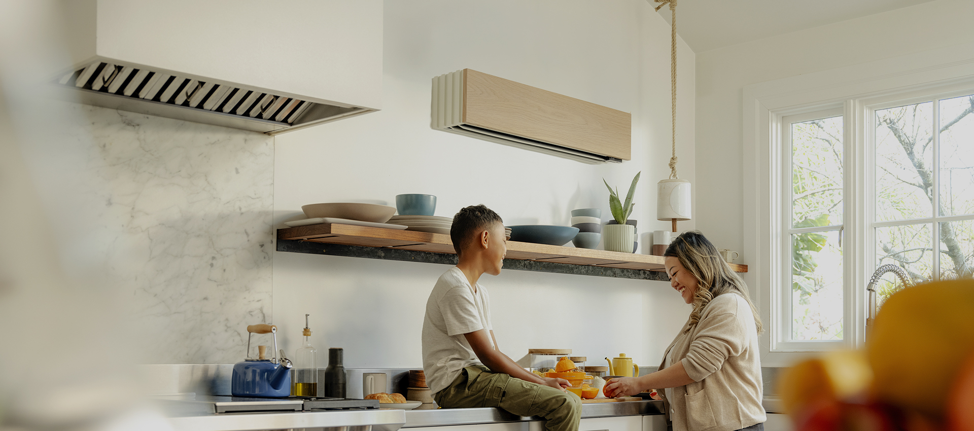 image.png A young boy sits on a kitchen counter talking to a woman who is preparing food. The modern kitchen has open shelves with dishes, a marble backsplash, and sunlight coming through a large window.