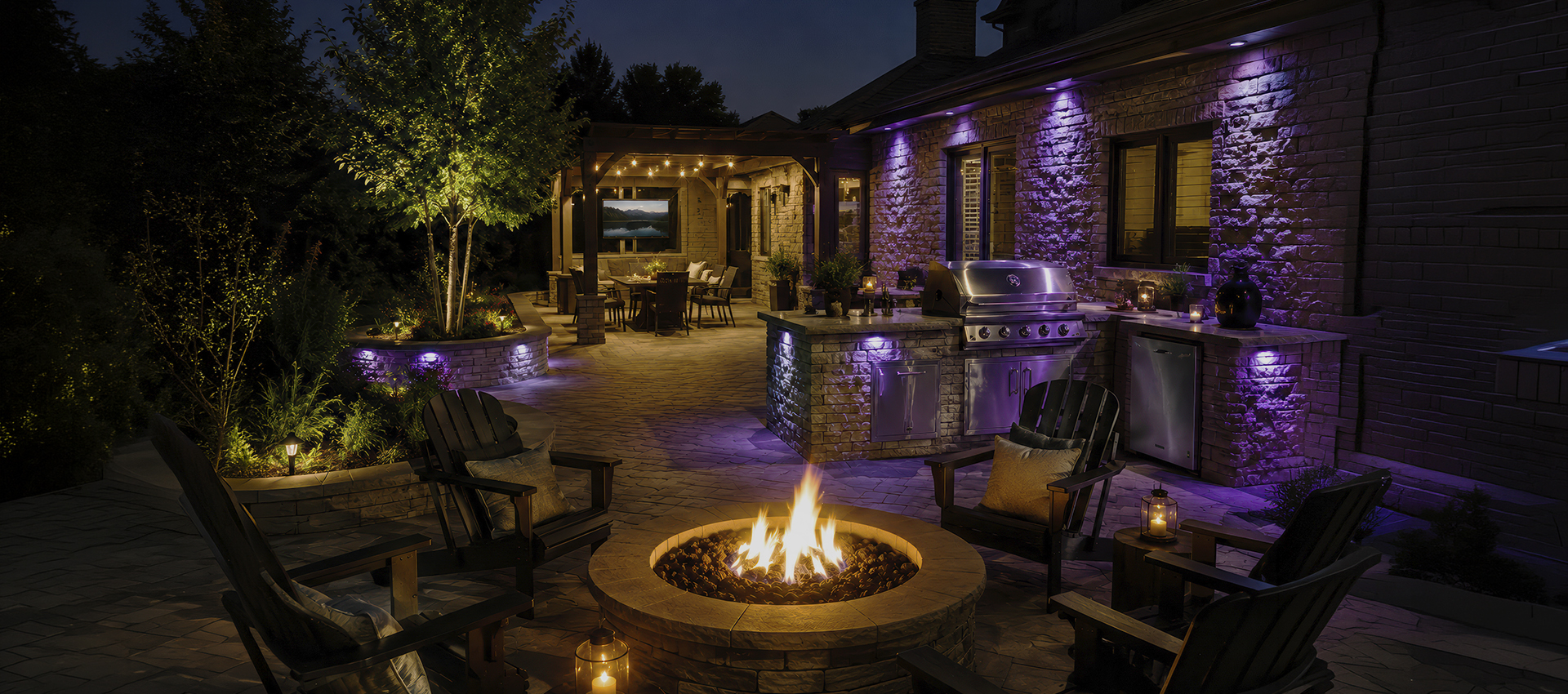 image.png A cozy backyard patio at night by Gandy Installations features a circular fire pit surrounded by chairs, an outdoor kitchen with a grill, dining area under a pergola, and purple accent lighting on stone walls and landscaping.