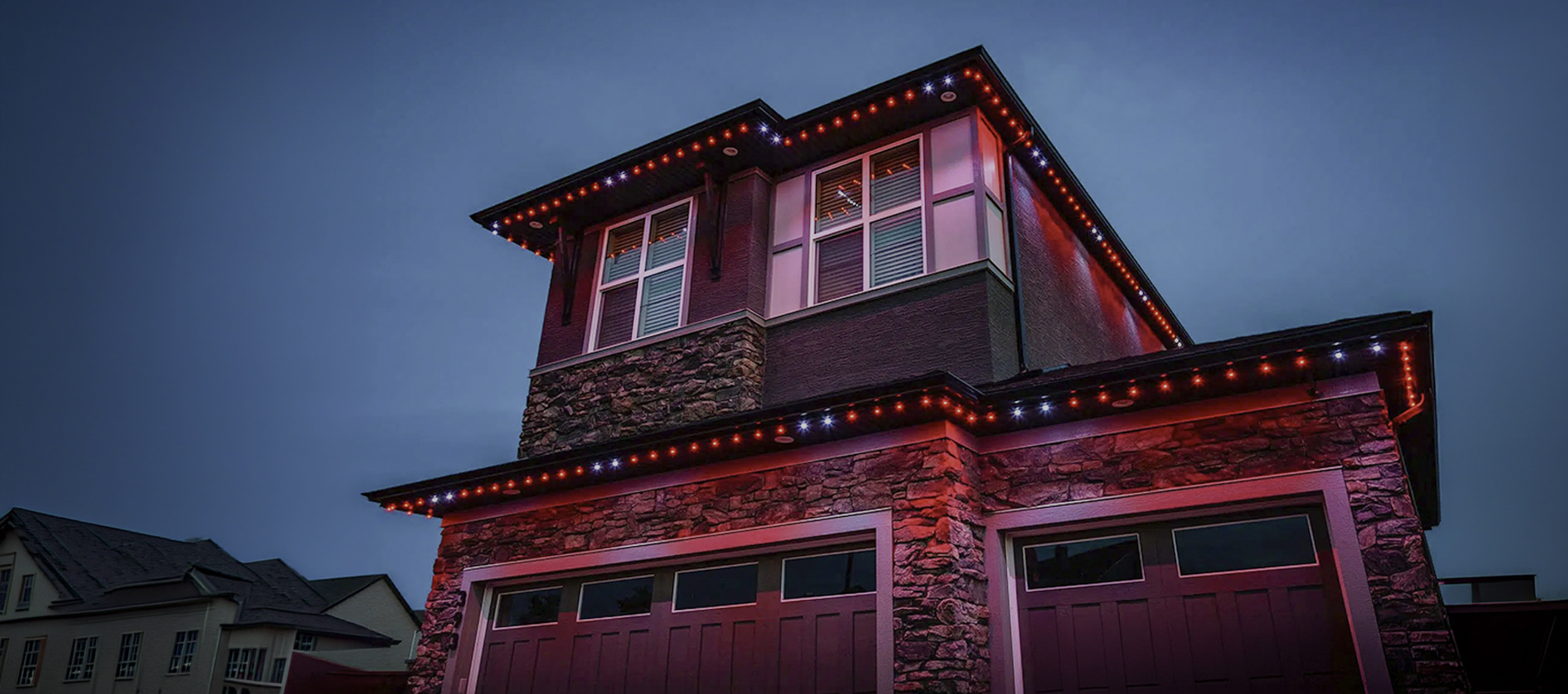 Two-story house at dusk with stone and stucco exterior, red and white lights outlining the roof and garage, creating a glowing effect against the darkening sky—a beautiful display expertly crafted by Gandy Installations.